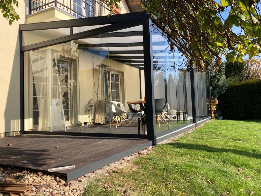 Zadaszony taras z metalową konstrukcją i szklanymi ścianami, widok z zewnątrz na drewniany podest i trawnik.