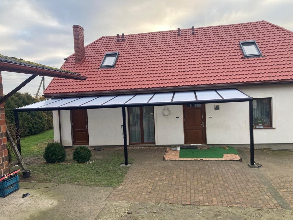 Metalowa pergola z transparentnym zadaszeniem przy białym domu z czerwoną dachówką i oknami dachowymi, widok z zewnątrz na kostkę brukową i niskie krzewy.