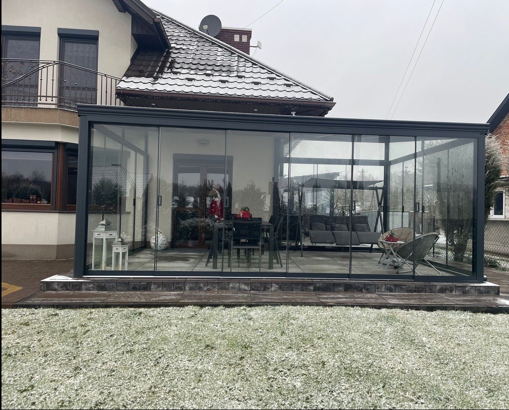 Szklana konstrukcja przylegająca do domu, prawdopodobnie oranżeria lub weranda, z metalową ramą, widoczna trawa pokryta szronem i meble ogrodowe wewnątrz