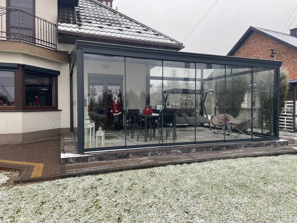 Zimowy ogród zimowy z ciemną ramą, przeszklony, z meblami wypoczynkowymi w środku, widok z zewnątrz na lekko ośnieżony trawnik.