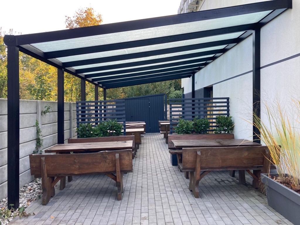Zadaszone patio z metalową konstrukcją, drewnianymi stołami i ławkami, oraz roślinami w donicach, przylegające do budynku z szarą elewacją.