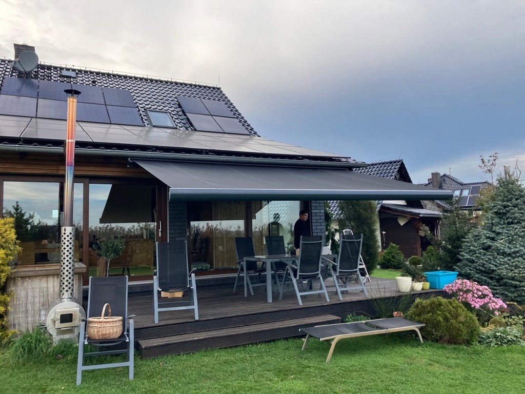 Zadaszony taras z wysuwaną markizą w kolorze grafitowym, metalowy komin pieca opalanego drewnem, panele słoneczne na dachu domu jednorodzinnego.