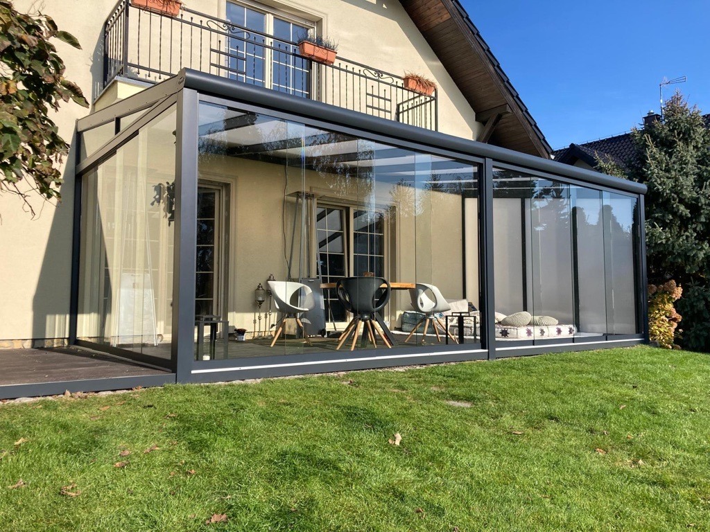 Szklana konstrukcja z czarną ramą przylegająca do domu, widok z zewnątrz na meble ogrodowe i poduszki wewnątrz, trawnik na pierwszym planie.