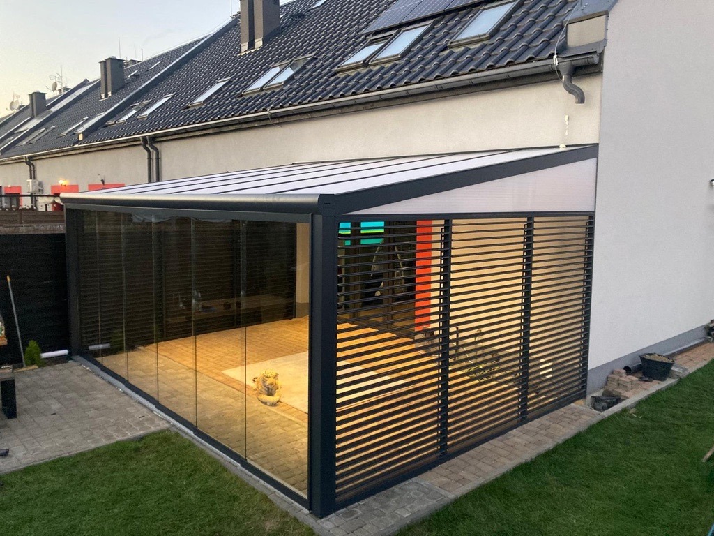 Nowoczesna pergola metalowa z lamelami i szklanymi ścianami, przylegająca do budynku mieszkalnego, widok z zewnątrz na ogród z trawnikiem i kostką brukową.