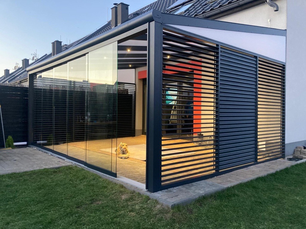 Nowoczesna konstrukcja z metalu i szkła, tworząca zadaszone patio z drewnianą podłogą, częściowo osłonięte żaluzjami, z figurką psa wewnątrz, na tle zadbanego trawnika.