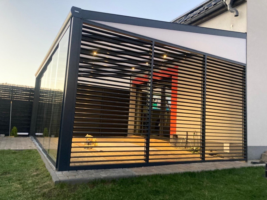 Nowoczesna konstrukcja z metalowych profili i szkła, tworząca zadaszone patio z oświetleniem punktowym, widok z zewnątrz na tle trawnika i domu.