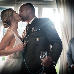 Studio Alternatywna - Pocałunek pary młodej w świetle okna; panna młoda w sukni z koralikami, pan młody w mundurze Wojska Polskiego, w tle mężczyzna w szarym garniturze.