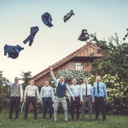 Studio Alternatywna - Grupowe zdjęcie uśmiechniętych mężczyzn w eleganckich strojach, rzucających marynarki w powietrze przed drewnianym domem z białymi kwiatami.