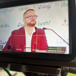 Studio Alternatywna - Ekran kamery Blackmagic Design pokazujący mężczyznę w czerwonej marynarce przemawiającego do mikrofonu na tle logotypów 'Dietly' i 'jush!', z widocznymi ustawieniami kamery i brakiem kart pamięci.