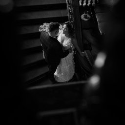 Studio Alternatywna - Czarno-białe zdjęcie pary młodej na schodach z ozdobną balustradą, panna młoda w okularach i sukni z aplikacjami kwiatowymi, pan młody w marynarce w kratę.