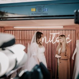 Studio Alternatywna - Panna młoda w koronkowej sukni i welonie rozmawia z konsultantką w salonie sukien ślubnych z neonowym napisem 'miłość' w tle, widok zza obiektywu kamery.