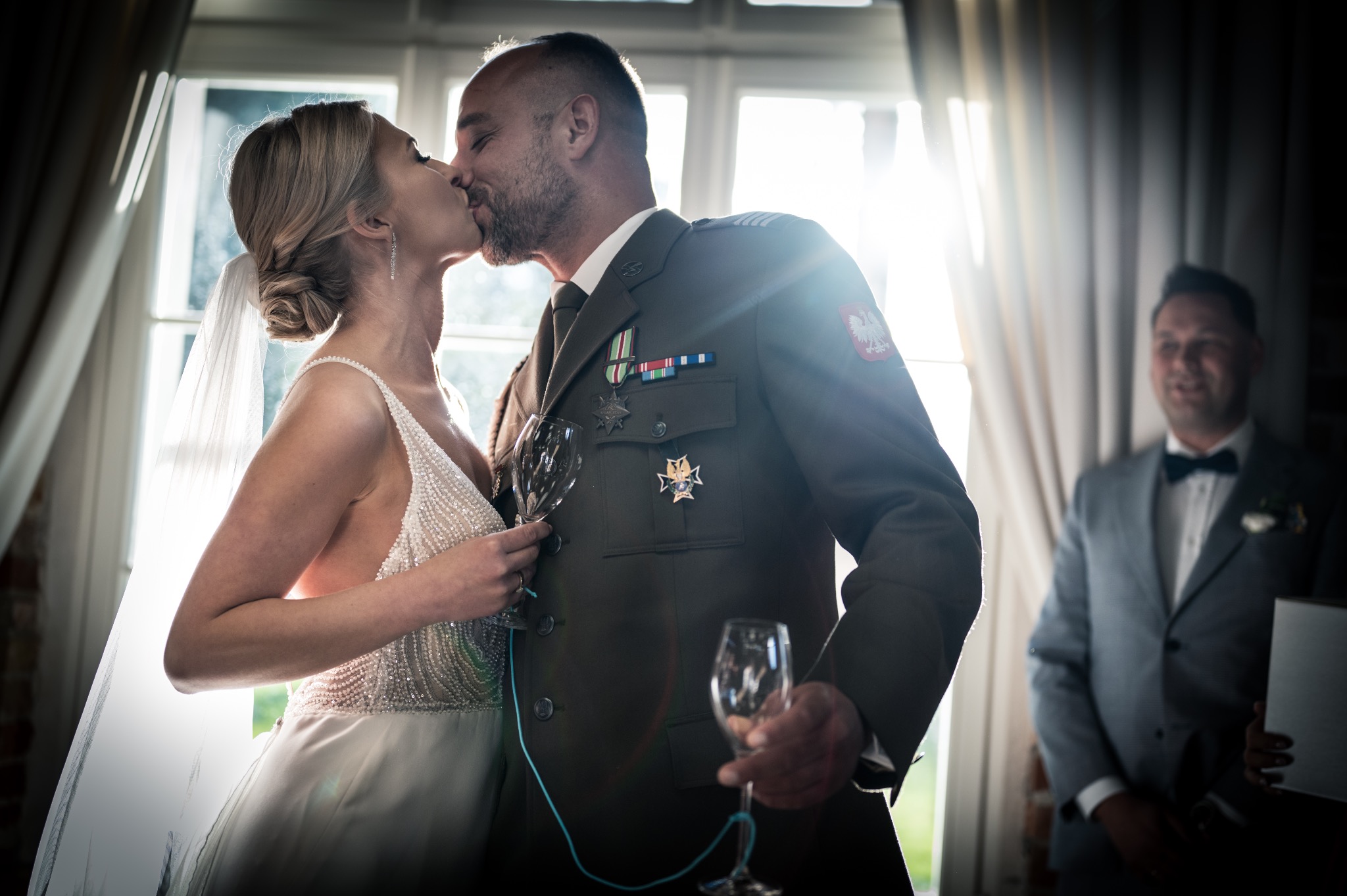 Pocałunek pary młodej w świetle okna; panna młoda w sukni z koralikami, pan młody w mundurze Wojska Polskiego, w tle mężczyzna w szarym garniturze.