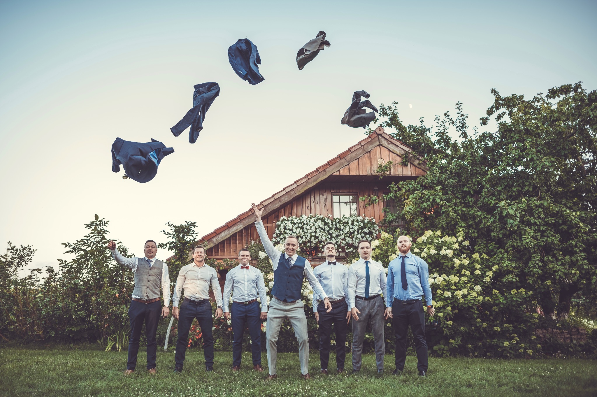 Grupowe zdjęcie uśmiechniętych mężczyzn w eleganckich strojach, rzucających marynarki w powietrze przed drewnianym domem z białymi kwiatami.