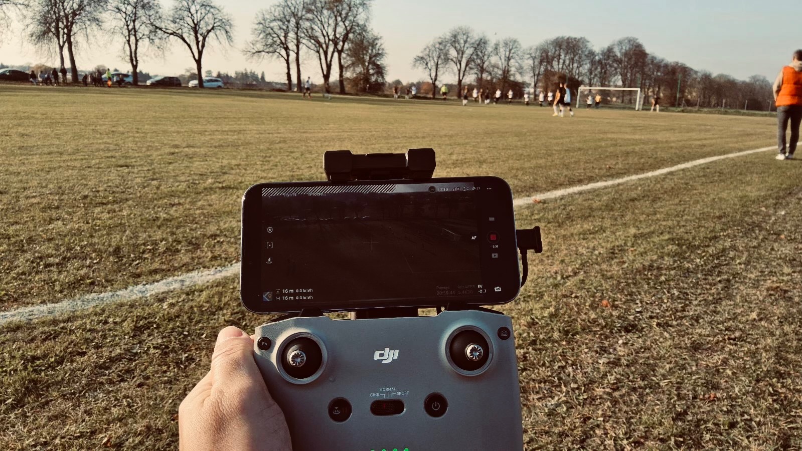 Operator drona z kontrolerem i smartfonem, na ekranie podgląd z kamery drona podczas filmowania meczu piłki nożnej na boisku.