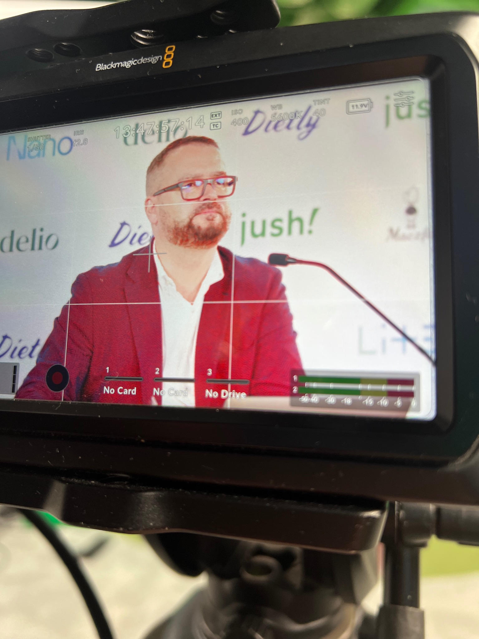 Ekran kamery Blackmagic Design pokazujący mężczyznę w czerwonej marynarce przemawiającego do mikrofonu na tle logotypów 'Dietly' i 'jush!', z widocznymi ustawieniami kamery i brakiem kart pamięci.