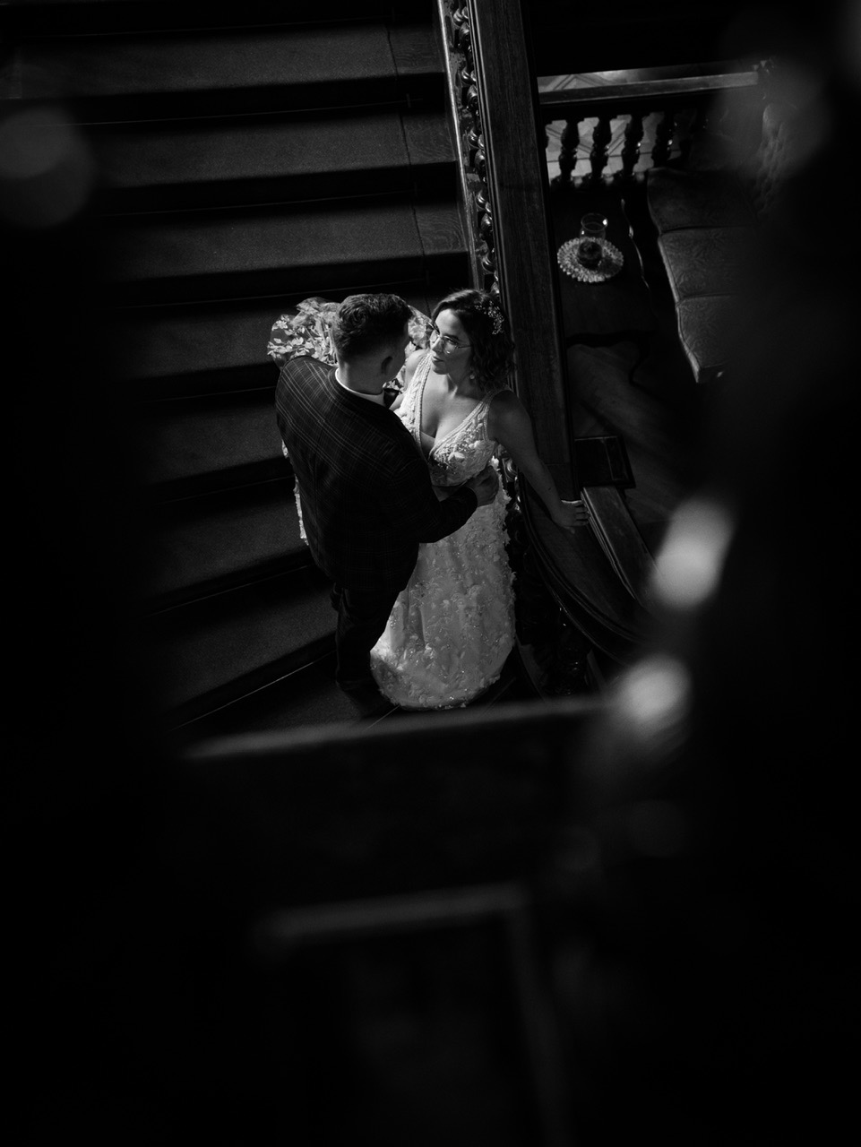 Czarno-białe zdjęcie pary młodej na schodach z ozdobną balustradą, panna młoda w okularach i sukni z aplikacjami kwiatowymi, pan młody w marynarce w kratę.