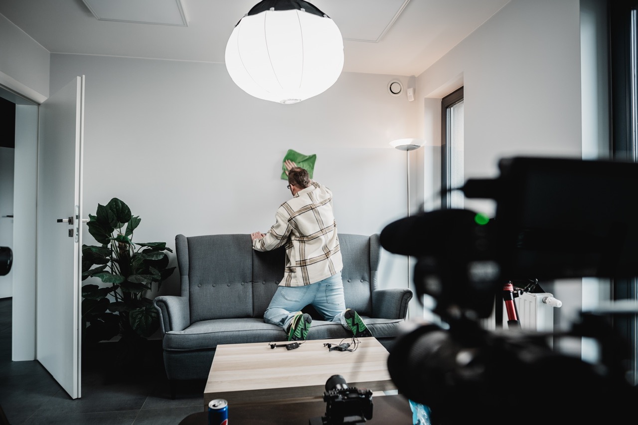 Mężczyzna w koszuli w kratę czyści ścianę w pokoju, opierając się o szarą sofę, widoczny zza kamery filmowej.