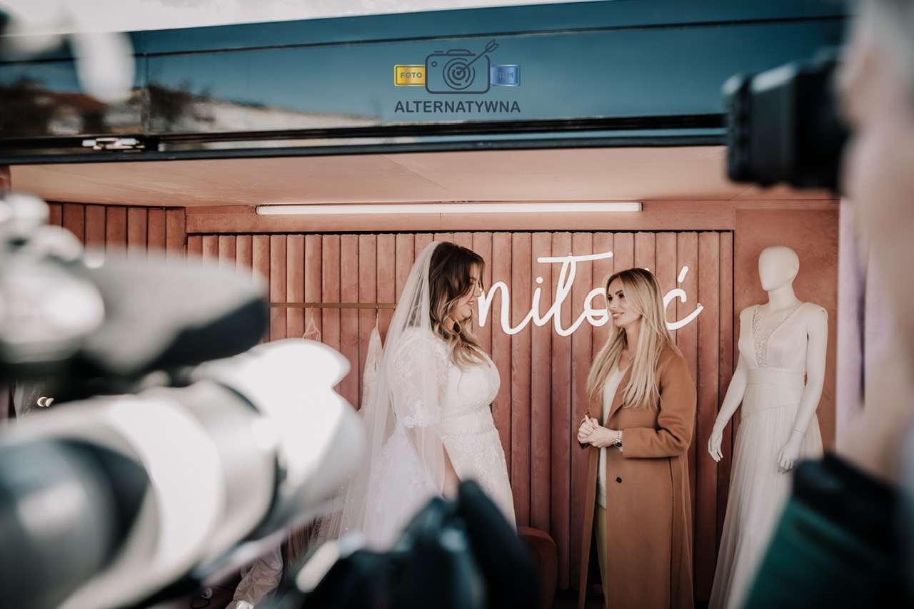 Panna młoda w koronkowej sukni i welonie rozmawia z konsultantką w salonie sukien ślubnych z neonowym napisem 'miłość' w tle, widok zza obiektywu kamery.