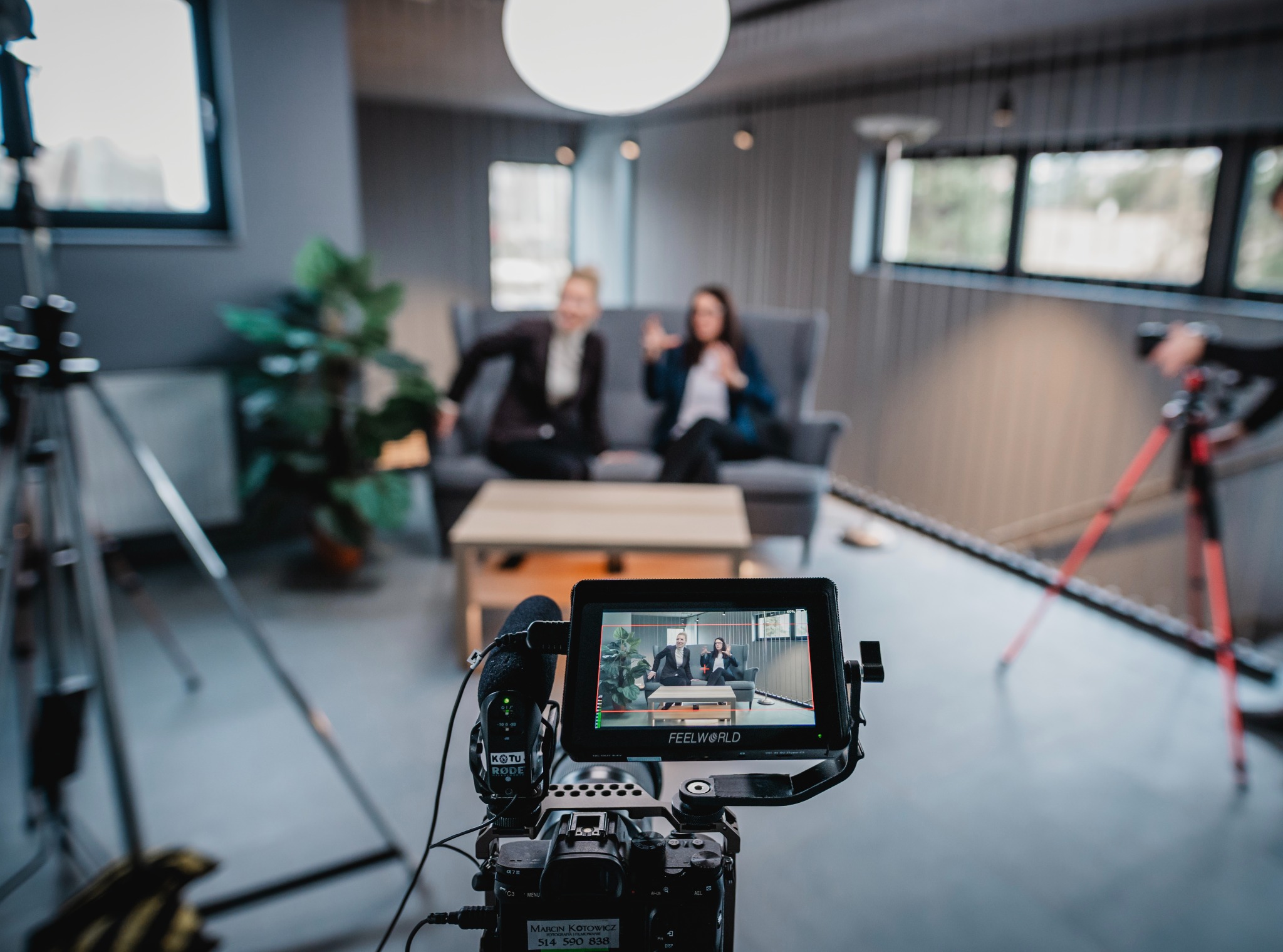 Ujęcie zza kamery, na ekranie monitora podgląd dwójki osób w garniturach siedzących na kanapie podczas nagrania w studio.