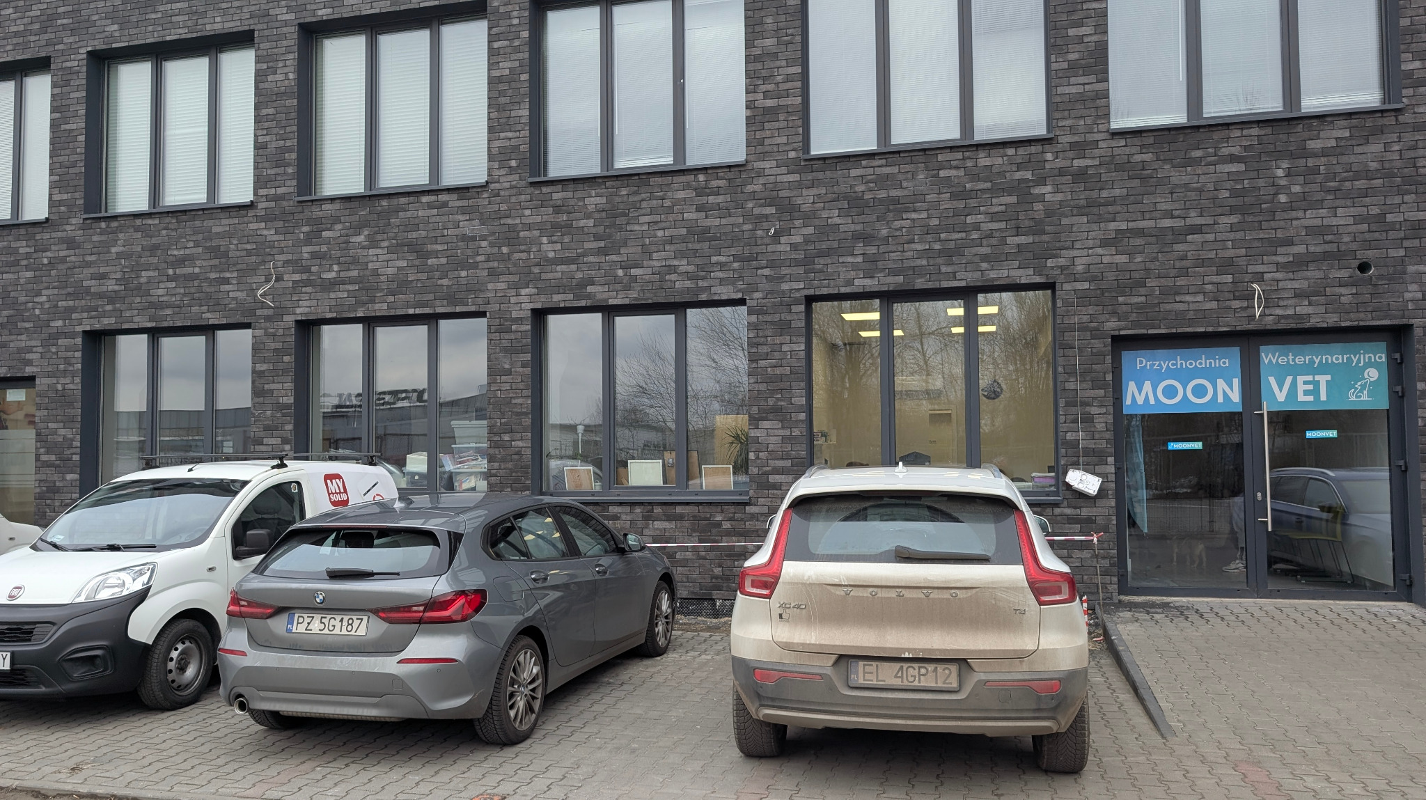 Nowoczesny budynek przychodni weterynaryjnej Moon Vet z szarym, ceglanym wykończeniem. Przed budynkiem zaparkowane samochody osobowe i dostawcze.