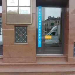 Wejście do budynku z napisem 'PSYCHOTESTY' na niebieskim pasku obok drzwi, numer 20 nad wejściem, tablica informacyjna z nazwami firm po lewej stronie, w odbiciu szyby widoczny samochód dostawczy...