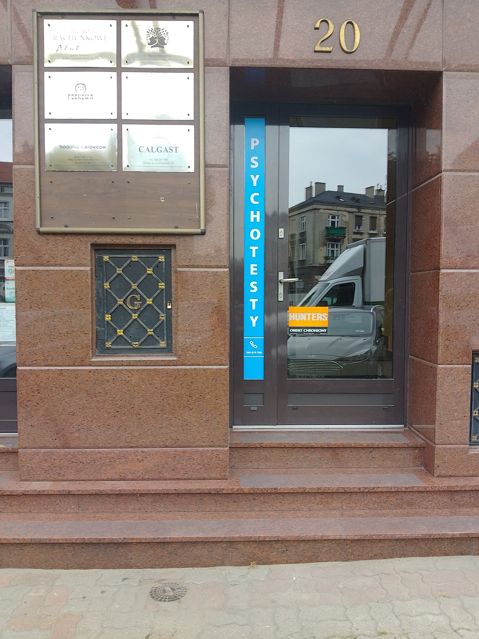 Wejście do budynku z napisem 'PSYCHOTESTY' na niebieskim pasku obok drzwi, numer 20 nad wejściem, tablica informacyjna z nazwami firm po lewej stronie, w odbiciu szyby widoczny samochód dostawczy...