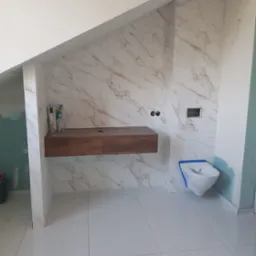 Łazienka w trakcie wykańczania: białe płytki imitujące marmur na ścianach, podwieszana szafka z ciemnego drewna, toaleta zabezpieczona niebieską taśmą, widoczne elementy instalacji.