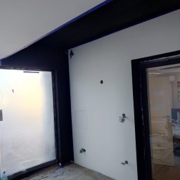 HRS interno sp.zoo - Białe ściany po malowaniu z czarnymi elementami wykończeniowymi, zabezpieczone folią okno i podłoga, widoczne ślady farby i taśmy malarskiej.
