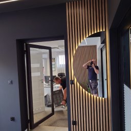 HRS interno sp.zoo - Nowoczesne wykończenie biura: drewniane lamele z podświetlanym lustrem, czarne ściany i przeszklone drzwi. Widoczny fragment podłogi zabezpieczony taśmą.