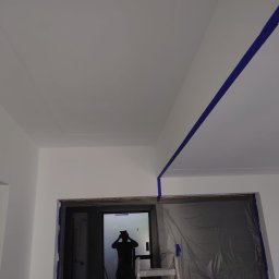 HRS interno sp.zoo - Białe ściany i sufit po malowaniu, zabezpieczone taśmą malarską, drabina w pokoju z oknem zabezpieczonym folią malarską. Widoczny cień osoby w oknie.