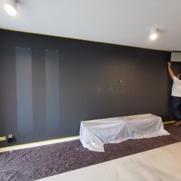 HRS interno sp.zoo - Malowanie ściany na ciemny kolor w pokoju. Mężczyzna maluje górną część ściany przy klimatyzatorze. Podłoga i meble zabezpieczone folią i taśmą malarską.