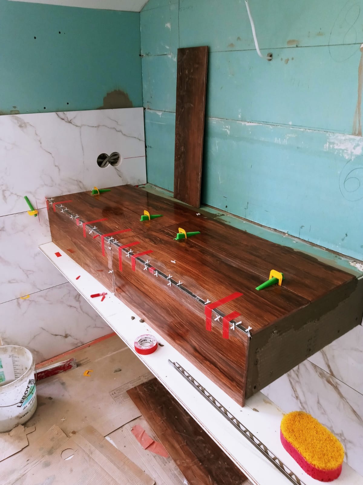 Montaż płytek imitujących drewno na obudowie w łazience, widoczne krzyżyki dystansowe, taśma malarska i poziomica, ściany w trakcie wykończenia.