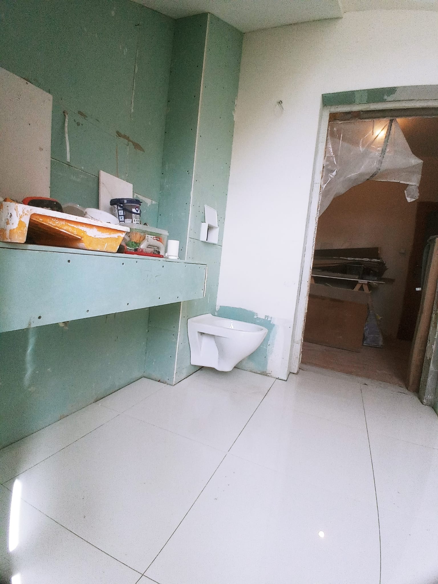 Łazienka w trakcie prac wykończeniowych: zamontowana toaleta wisząca, ściany przygotowane pod płytki gipsowo-kartonowe, widoczne narzędzia i materiały budowlane, białe płytki podłogowe.