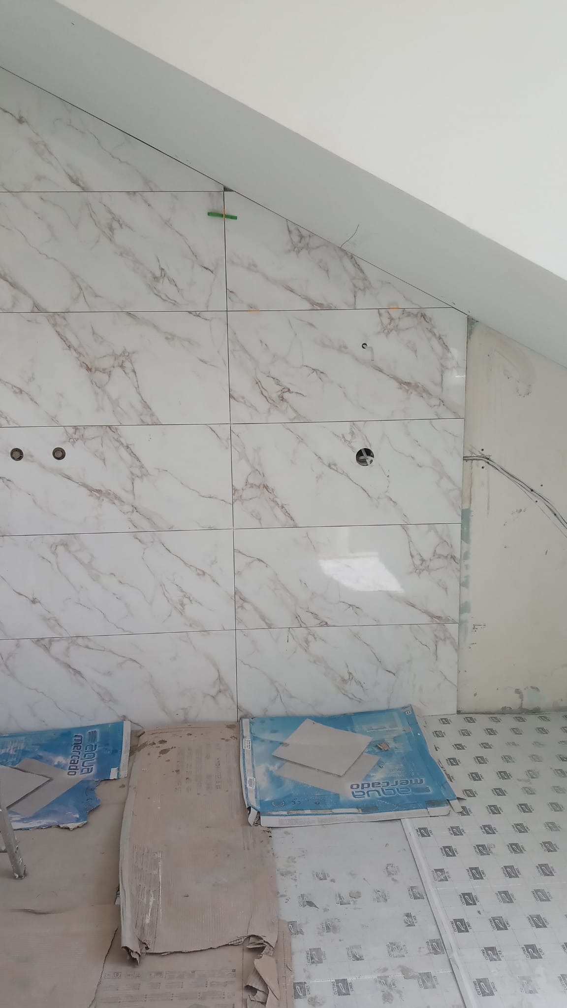 Układanie płytek imitujących marmur na ścianie ze skosem, widoczne paczki płytek na podłodze i zielone krzyżyki dystansowe między płytkami.