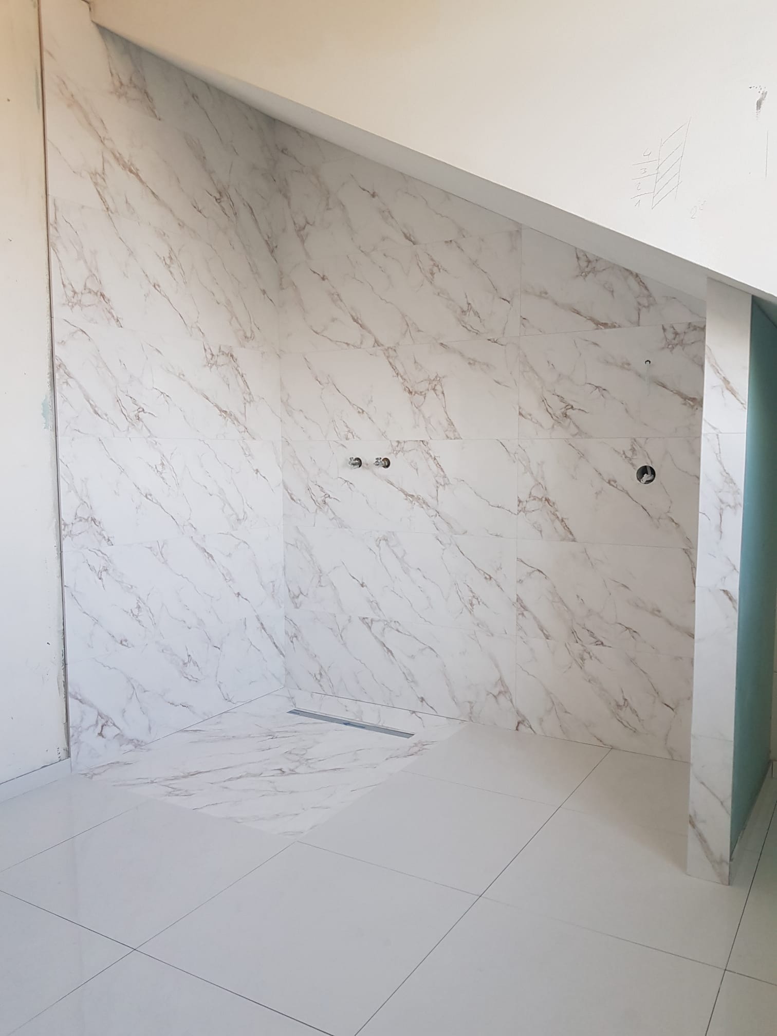 Płytki imitujące marmur na ścianach i podłodze w nowoczesnej łazience z odpływem liniowym, widoczne przygotowane otwory na armaturę.