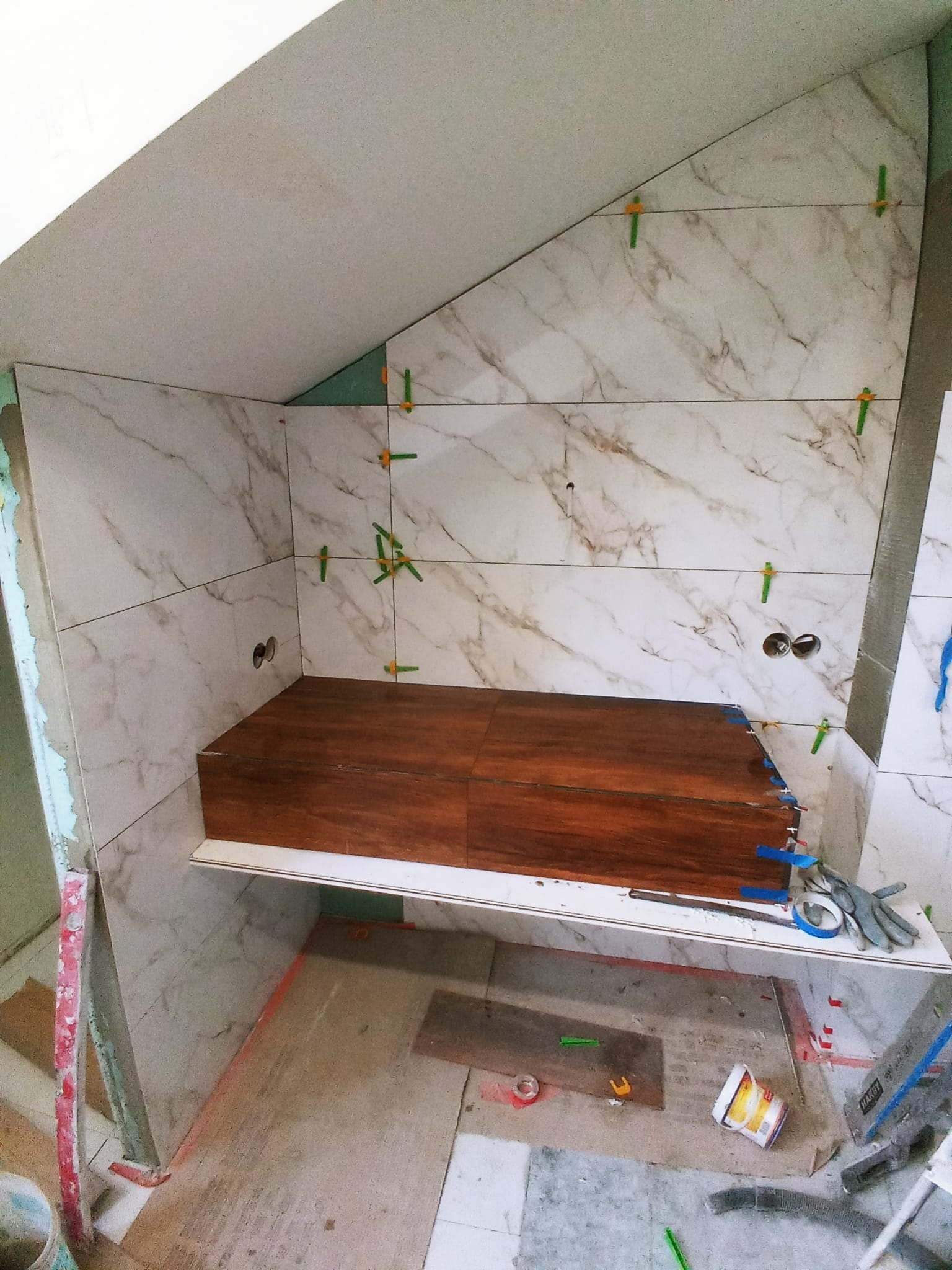 Wnęka w trakcie wykańczania płytkami imitującymi marmur, widoczne zielone krzyżyki dystansowe, drewniana półka z niedokończonymi brzegami, narzędzia i akcesoria glazurnicze w tle.