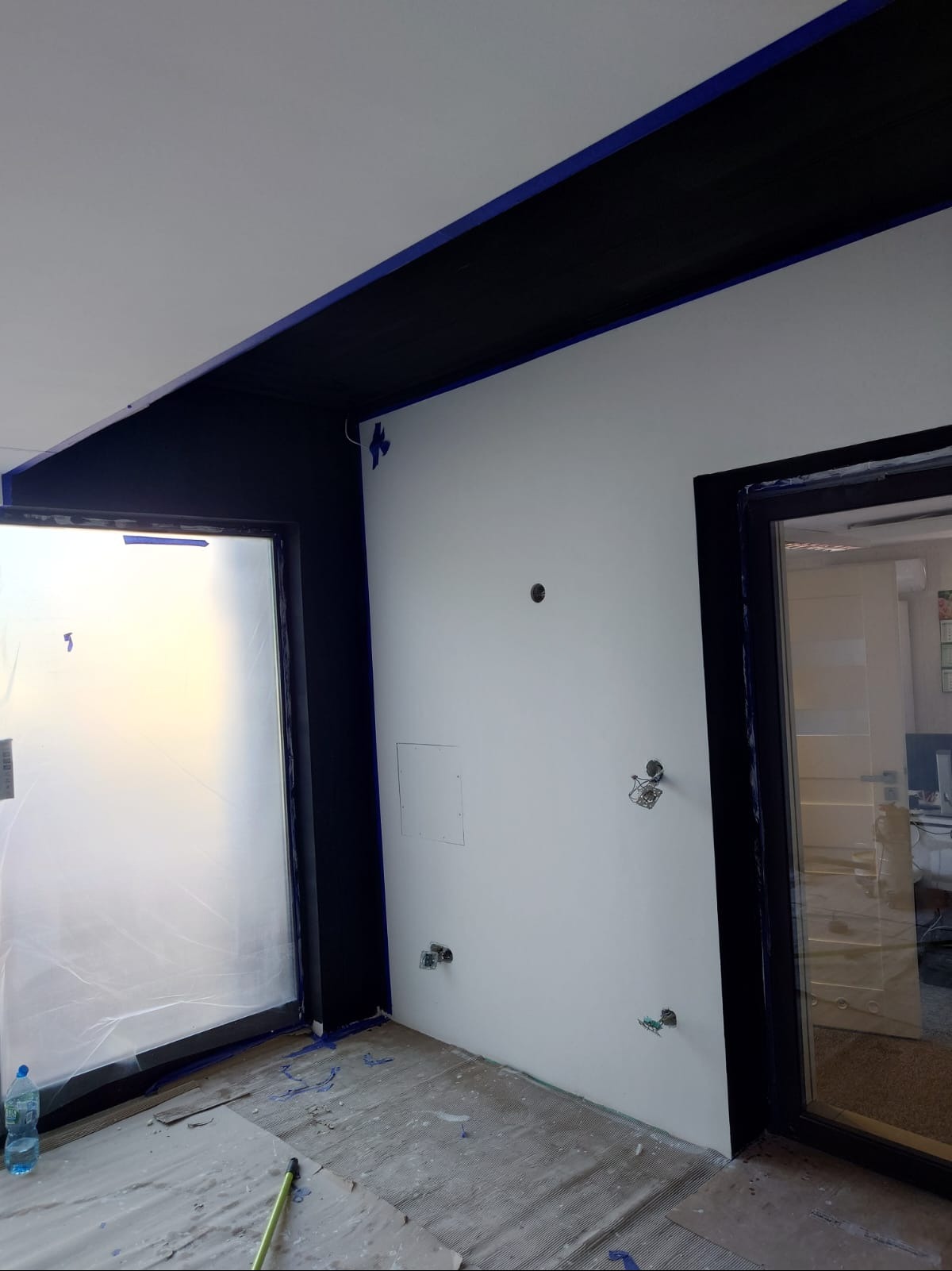 Białe ściany po malowaniu z czarnymi elementami wykończeniowymi, zabezpieczone folią okno i podłoga, widoczne ślady farby i taśmy malarskiej.