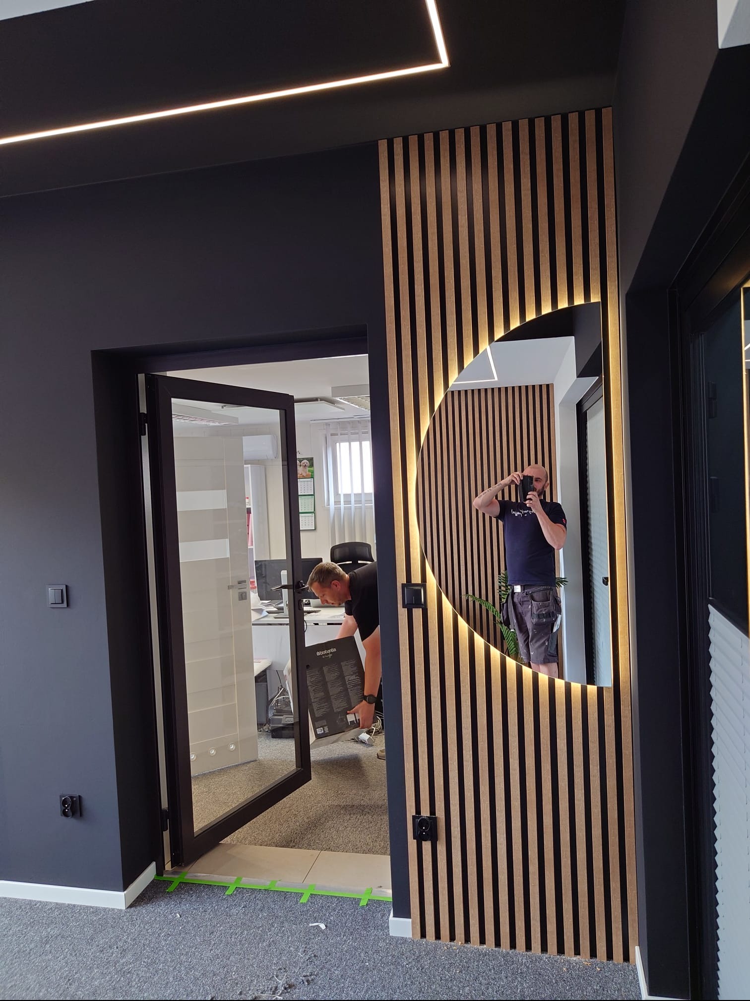 Nowoczesne wykończenie biura: drewniane lamele z podświetlanym lustrem, czarne ściany i przeszklone drzwi. Widoczny fragment podłogi zabezpieczony taśmą.