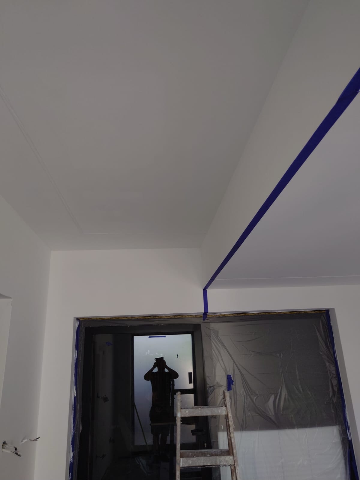 Białe ściany i sufit po malowaniu, zabezpieczone taśmą malarską, drabina w pokoju z oknem zabezpieczonym folią malarską. Widoczny cień osoby w oknie.