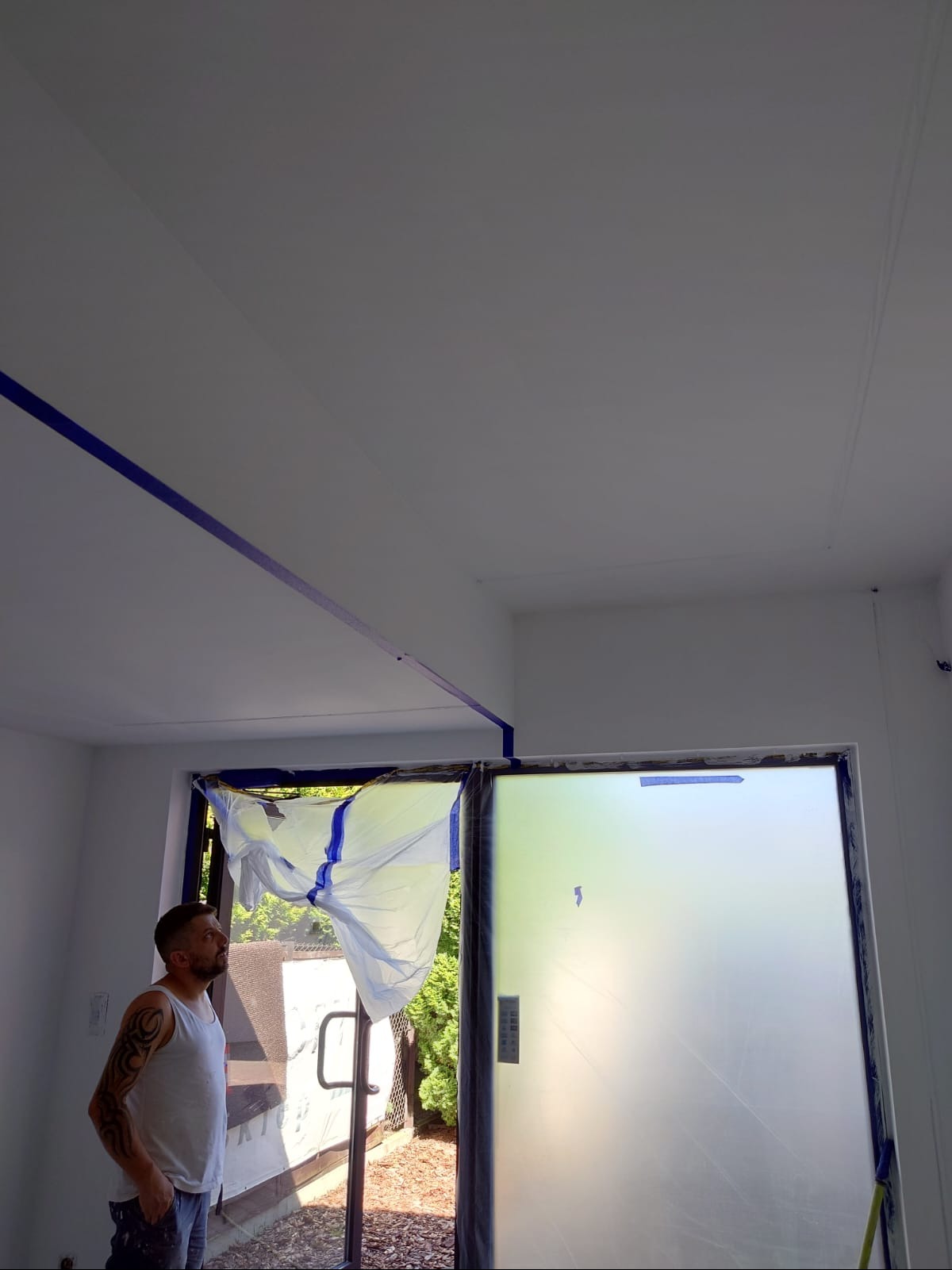 Przygotowania do malowania: Mężczyzna w białym podkoszulku patrzy na sufit zabezpieczony taśmą malarską, okno osłonięte folią. Widoczne drzewa za oknem.