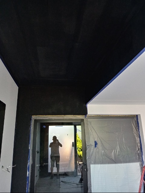 Częściowo pomalowane na czarno wnętrze z zabezpieczonymi folią drzwiami. Widoczny fragment białej ściany i sufitu. W tle osoba robiąca zdjęcie.