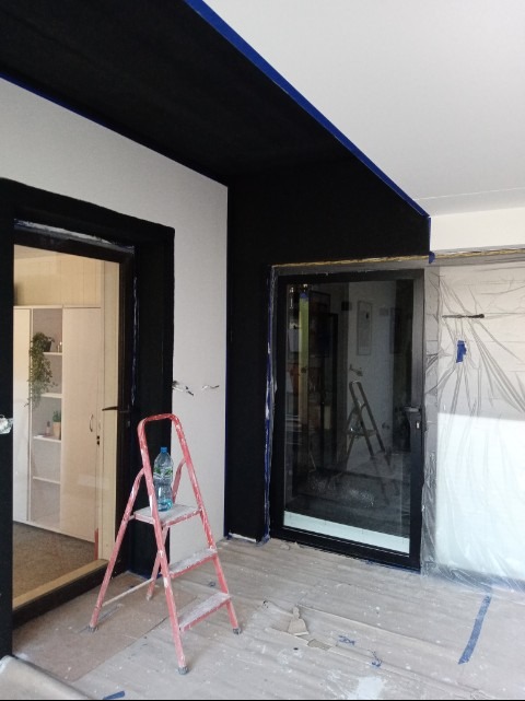 Wnętrze w trakcie malowania: czarne ściany i sufit, białe fragmenty ścian, zabezpieczone okno folią, drabina z butelką wody. Widoczne ślady farby na podłodze.