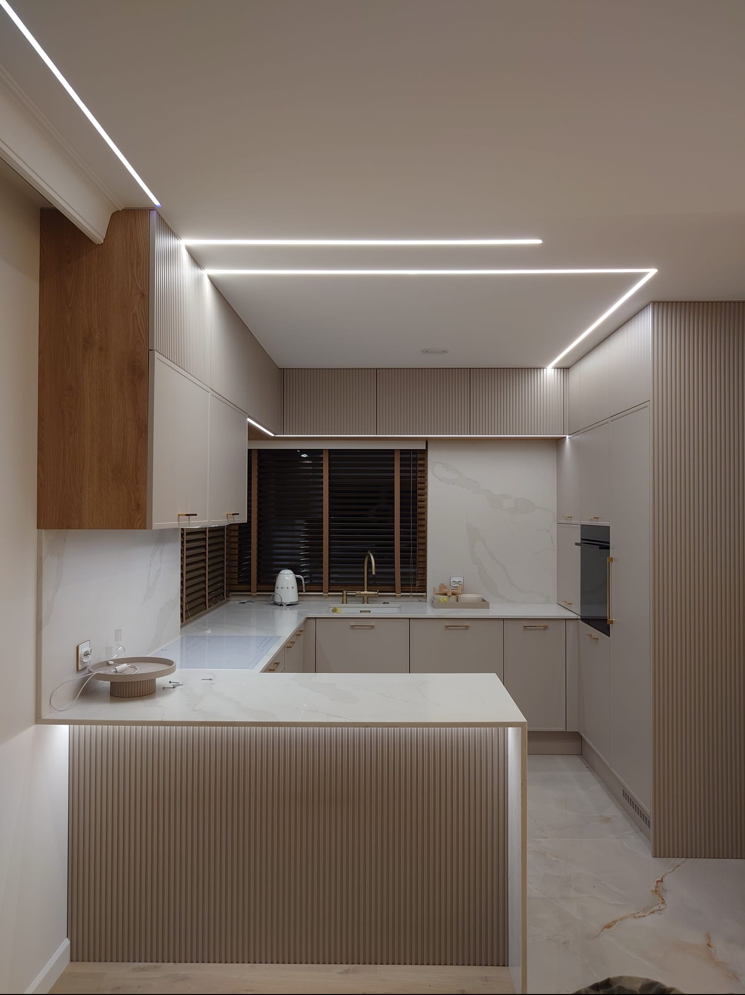 Nowoczesna kuchnia w beżach z marmurowym blatem i oświetleniem LED. Ryflowane fronty szafek i wyspa kuchenna. Minimalistyczny design z elementami drewna.