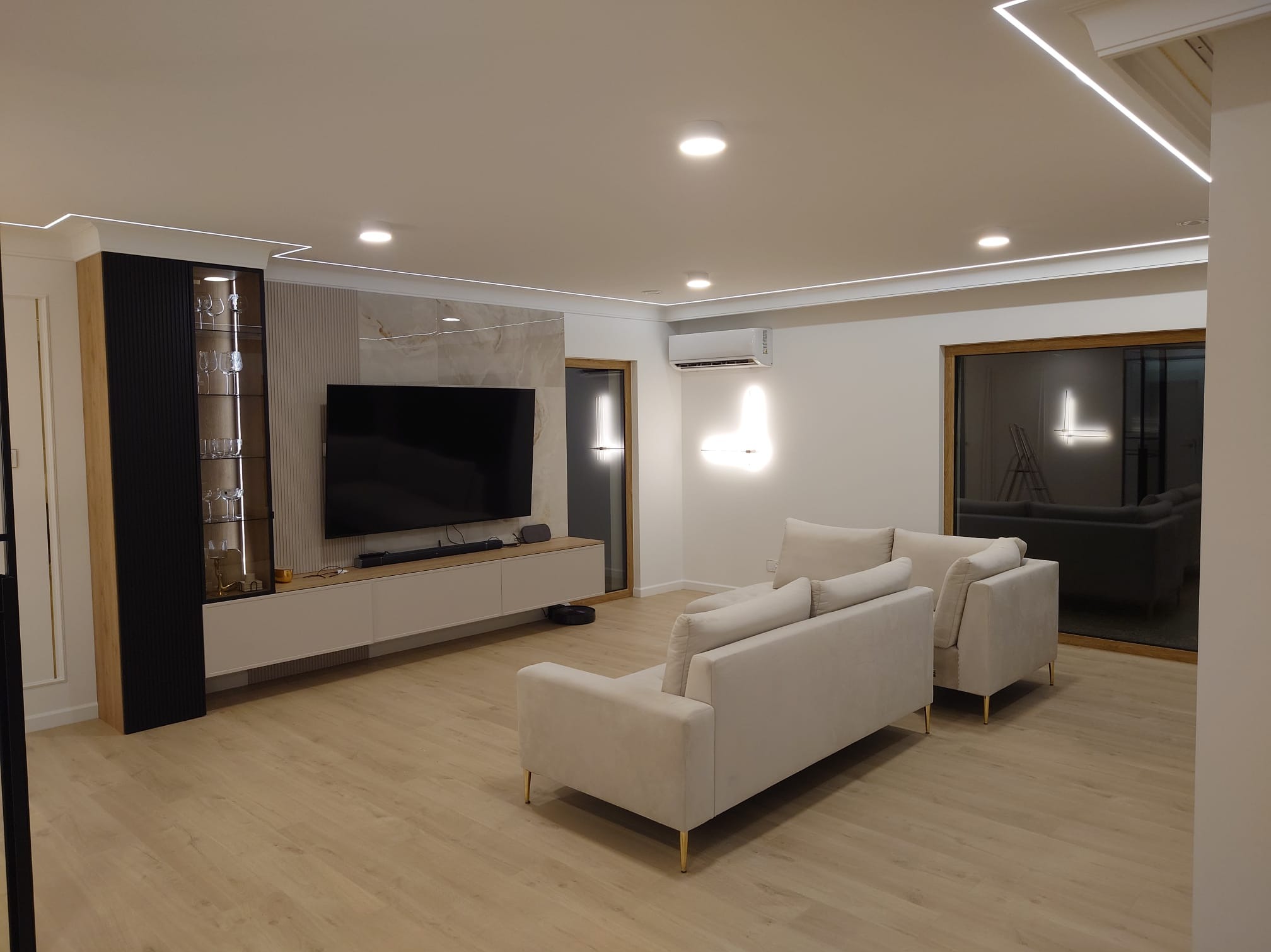 Nowoczesny salon z jasną podłogą, beżową sofą i podświetleniem LED na suficie. Widoczna szafka RTV z telewizorem i przeszklona witryna. Minimalistyczny design.