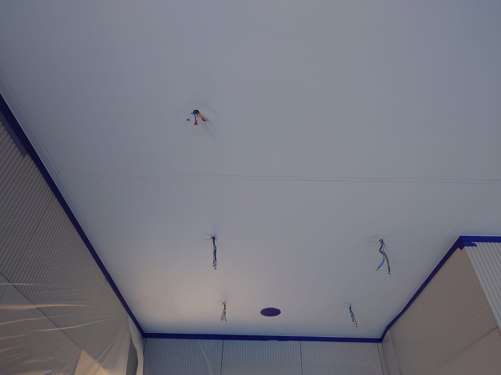 Biały sufit po malowaniu z widocznymi przewodami elektrycznymi i taśmą malarską zabezpieczającą ściany. Widoczne otwory na oświetlenie.