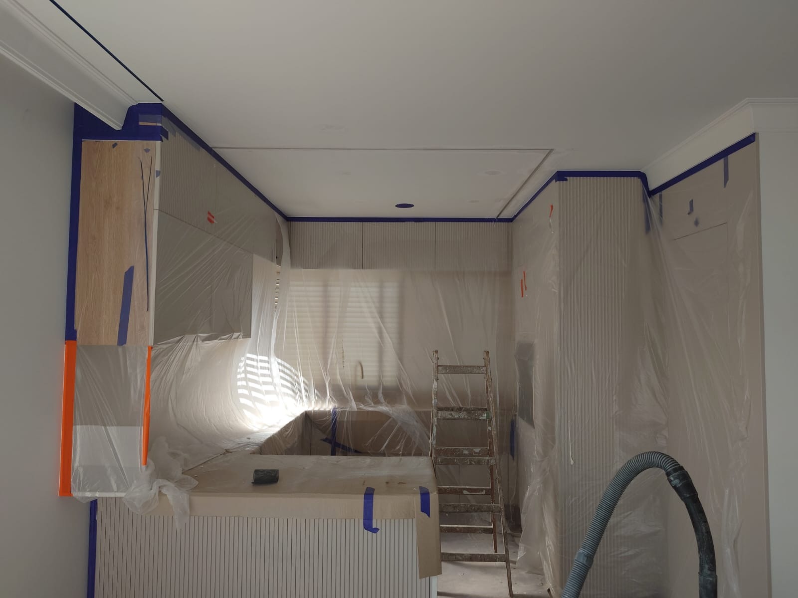 Kuchnia zabezpieczona folią i taśmą malarską przed malowaniem. Widoczna drabina i odkurzacz. Przygotowanie do prac malarskich, ochrona mebli i powierzchni.