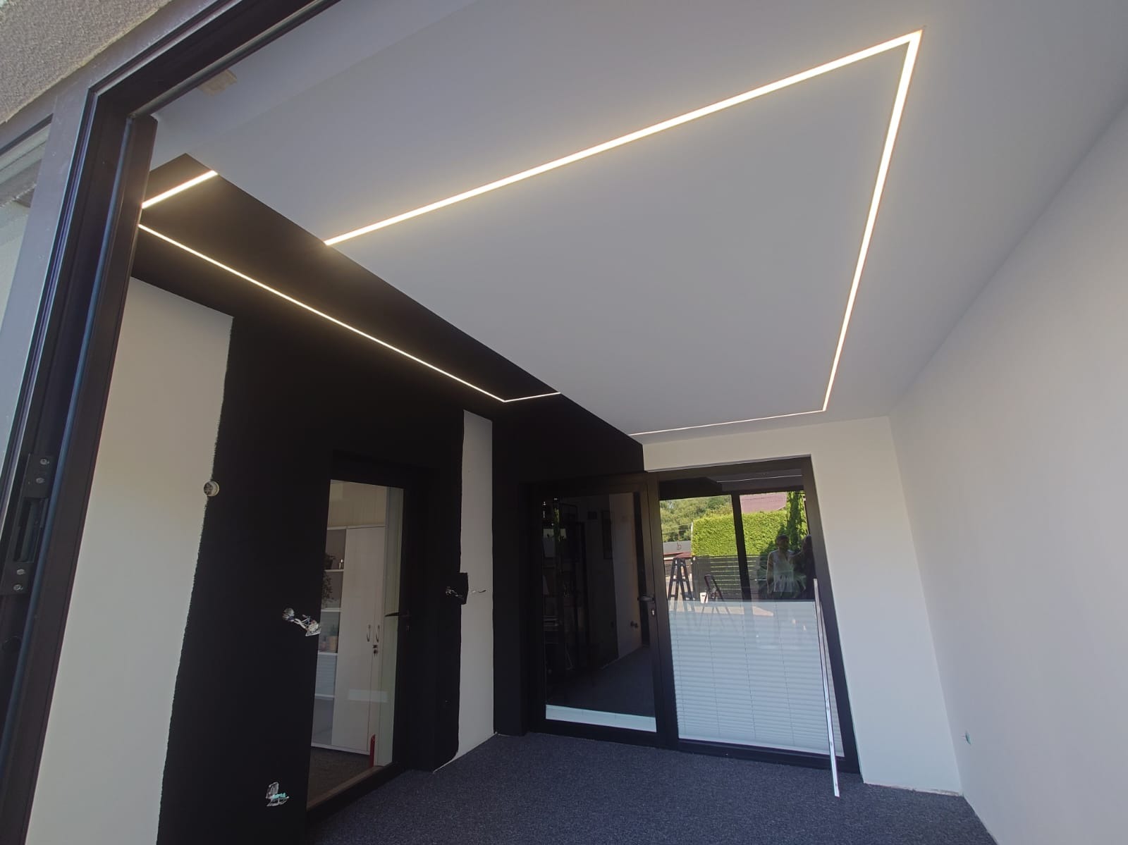 Nowoczesne oświetlenie LED w kształcie linii i trójkąta na suficie, czarno-białe ściany, szara podłoga, widok na zewnątrz przez szklane drzwi.