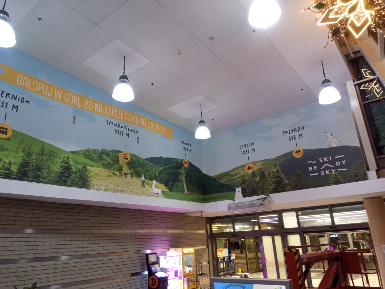 Wnętrze z muralem przedstawiającym górski krajobraz z nazwami szczytów i wysokościami, oświetlone lampami. Widoczne elementy architektoniczne i dekoracje.