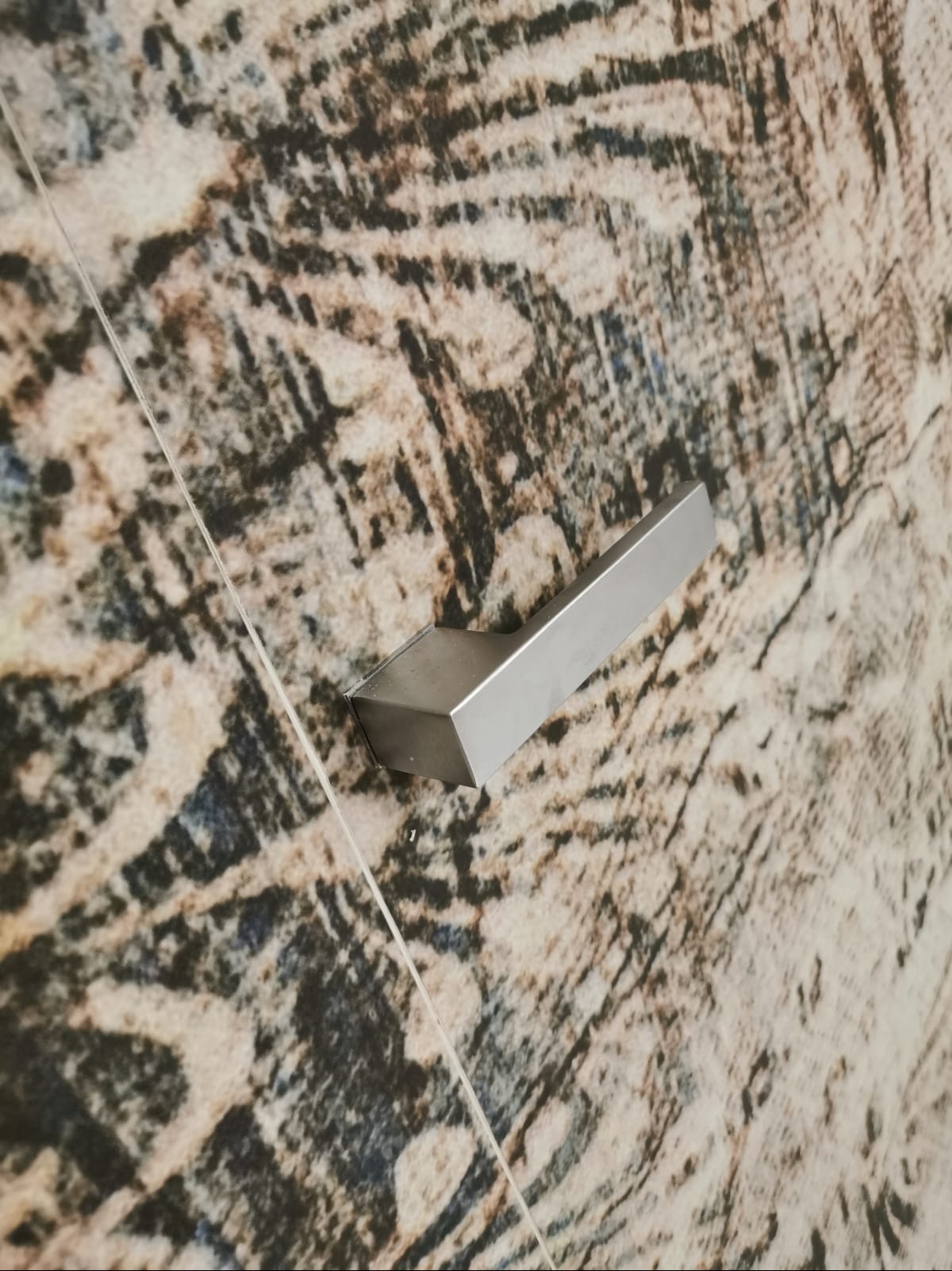 Szczegółowe ujęcie chromowanej klamki na tle ściany z wzorem imitującym naturalny kamień. Widoczna fuga między płytkami. Minimalistyczny design.