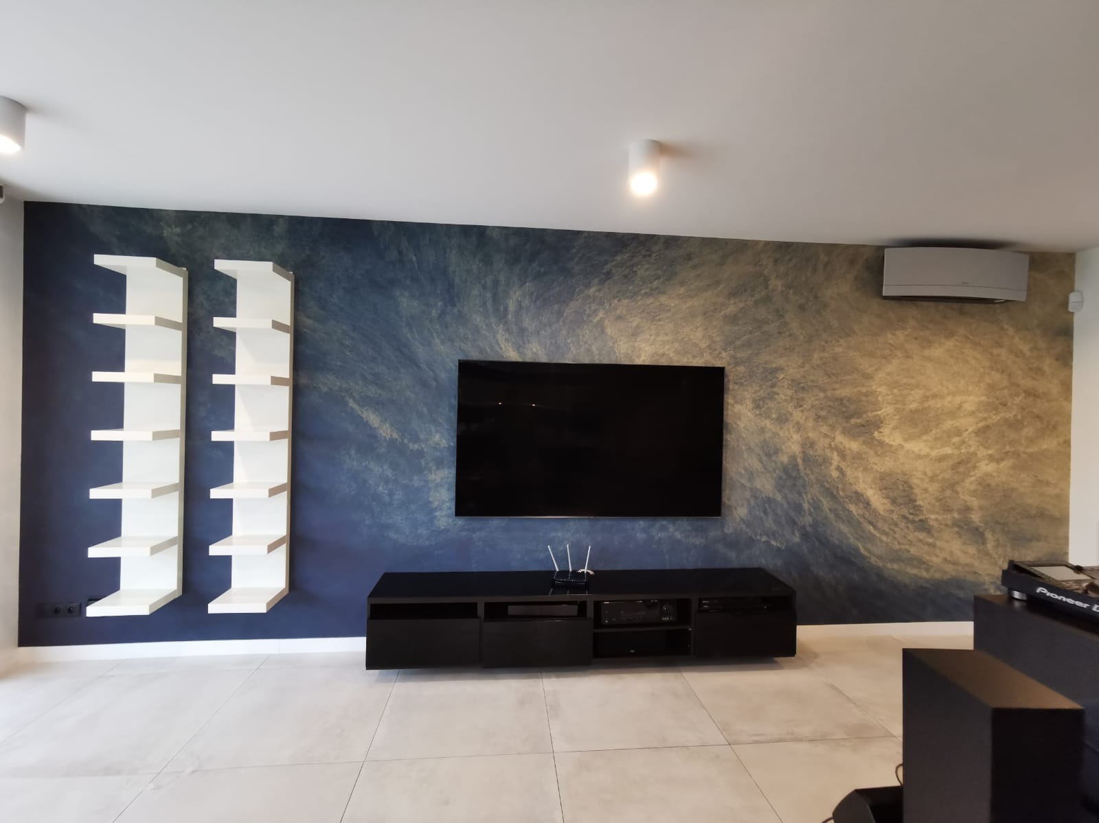 Ściana w salonie z dekoracyjnym malowaniem w odcieniach niebieskiego i beżu, z wiszącymi białymi półkami i czarną szafką RTV. Nowoczesny design, klimatyczne oświetlenie.